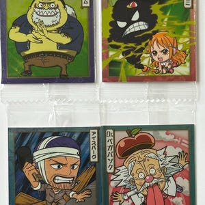 May include: Four colorful square cards featuring characters from the anime One Piece. The cards feature a yellow character with a beard, a black cloud with a face, a character with a bandage on his head, and a character with a red apple on his head.