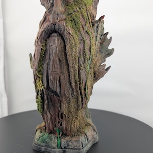 May include: A detailed sculpture of a tree-like figure with a face, beard, and moss accents. The figure is primarily brown with green moss and a textured surface. The sculpture is on a black surface.