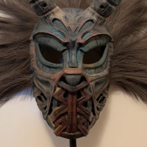 Könnte beinhalten: Eine detaillierte Maske mit großen, gebogenen Hörnern und einer strukturierten Oberfläche. Die Maske weist eine Kombination aus blauen, braunen und roten Farbtönen mit aufwendigen Schnitzereien auf. Graues Kunstfell umgibt die Maske.