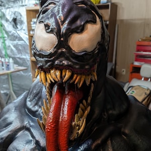 Puede incluir: Una escultura detallada del personaje Venom de Marvel Comics. El busto presenta una combinación de colores negro y azul oscuro con ojos blancos, dientes afilados y una larga lengua roja. La escultura tiene una apariencia texturizada.
