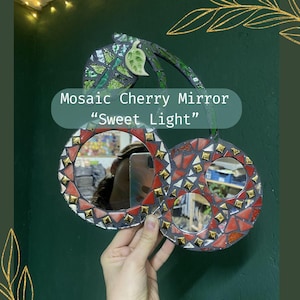 Könnte beinhalten: Mosaik-Kirschspiegel mit roten, goldenen und grünen Mosaikfliesen. Der Spiegel hat die Form von Kirschen, mit dem Text "Mosaic Cherry Mirror 'Sweet Light'" auf dem Bild. Der Spiegel wird von einer Hand gehalten.