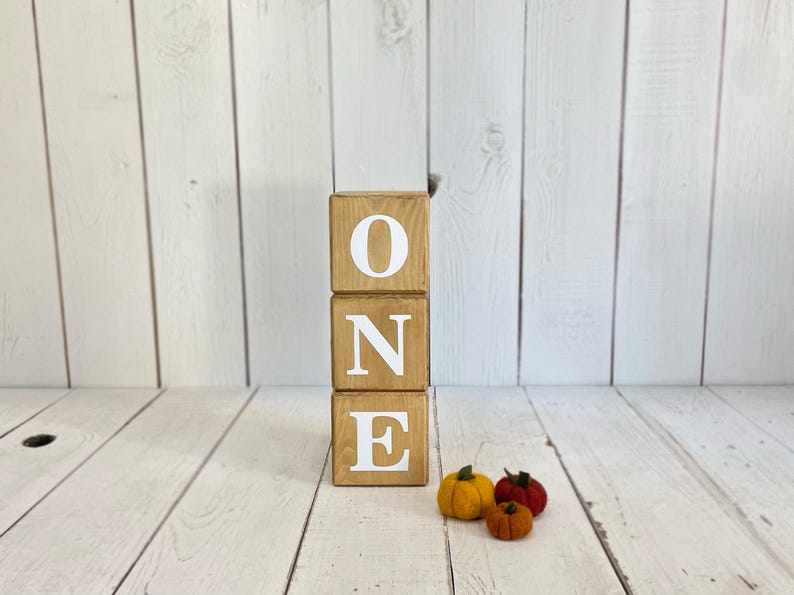 Puede incluir: Bloques de madera que deletrean la palabra "ONE" en letras blancas. Los bloques est&aacute;n apilados uno encima del otro. Hay tres peque&ntilde;as calabazas de fieltro delante de los bloques.