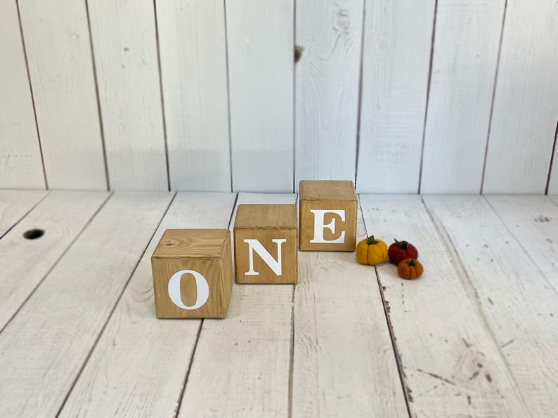 Puede incluir: Bloques de madera que deletrean la palabra "ONE" en letras blancas. Los bloques est&aacute;n dispuestos en l&iacute;nea sobre una superficie de madera blanca. Hay tres peque&ntilde;as calabazas de fieltro en el fondo.