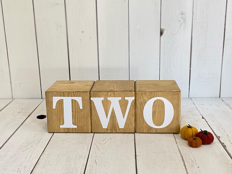 May include: Three wooden blocks spelling out the word "TWO" in white letters. The blocks are arranged in a row on a white wooden surface. There are three small felt pumpkins in the foreground.