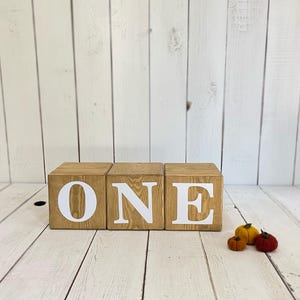 May include: Three wooden blocks spelling out the word "ONE" in white letters. The blocks are stacked on a white wood background.