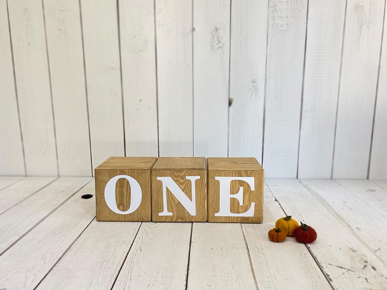 May include: Three wooden blocks spelling out the word "ONE" in white letters. The blocks are stacked on a white wood background.