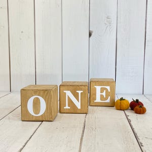 Puede incluir: Bloques de madera que deletrean la palabra "ONE" en letras blancas. Los bloques est&aacute;n dispuestos en una fila sobre una superficie de madera blanca. Hay tres peque&ntilde;as calabazas de fieltro de color naranja y amarillo en primer plano.
