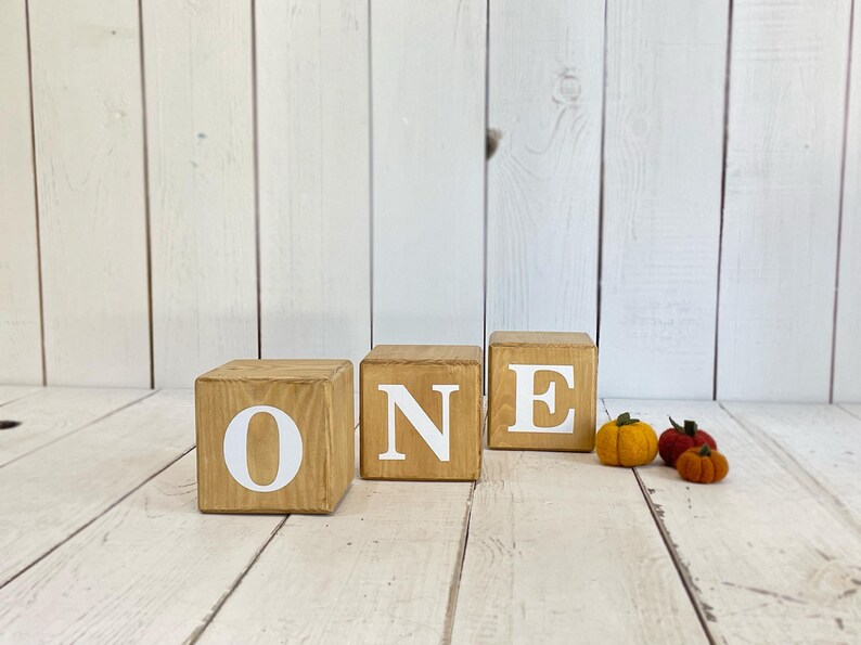 Puede incluir: Bloques de madera que deletrean la palabra "ONE" en letras blancas. Los bloques est&aacute;n dispuestos en una fila sobre una superficie de madera blanca. Hay tres peque&ntilde;as calabazas de fieltro de color naranja y amarillo en primer plano.