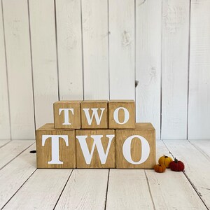 May include: Wooden blocks spelling out the word "TWO" in white letters. The blocks are stacked in two rows, with the top row spelling "TWO" and the bottom row spelling "TWO".