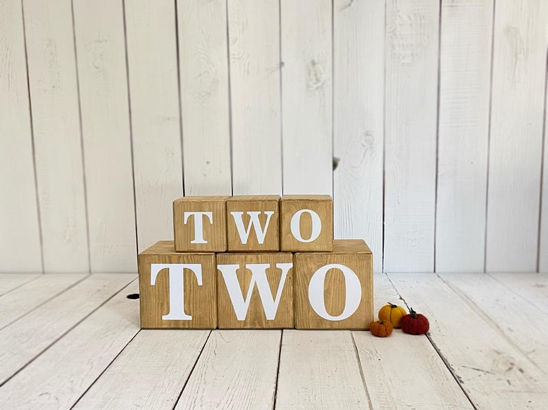 May include: Wooden blocks spelling out the word "TWO" in white letters. The blocks are stacked in two rows, with the top row spelling "TWO" and the bottom row spelling "TWO".
