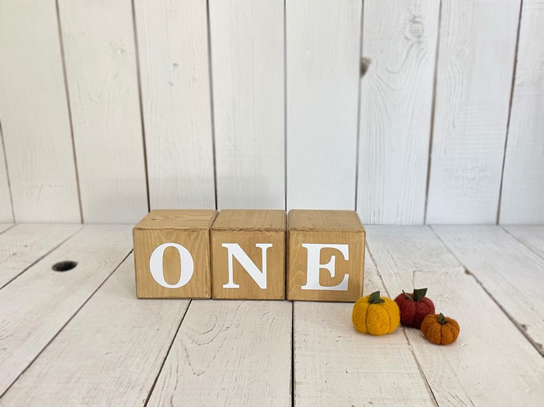 Puede incluir: Tres bloques de madera que deletrean la palabra "ONE" en letras blancas. Los bloques est&aacute;n sobre una superficie de madera blanca. Hay tres peque&ntilde;as calabazas de fieltro de color naranja y amarillo en primer plano.