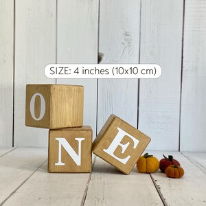 Puede incluir: Tres bloques de madera que deletrean la palabra "ONE" en letras blancas. Los bloques miden 4 pulgadas (10x10 cm) y est&aacute;n sobre un fondo de madera blanca. Hay tres peque&ntilde;as calabazas de fieltro naranja en primer plano.