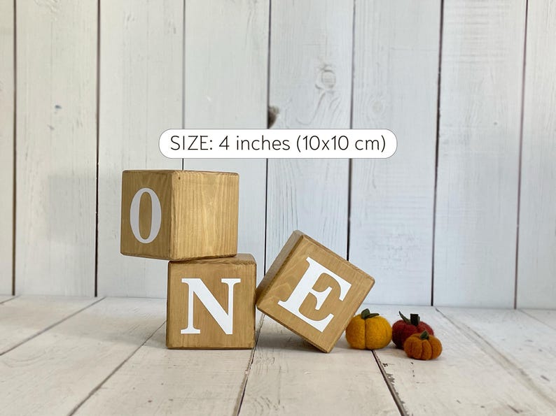 Puede incluir: Tres bloques de madera que deletrean la palabra "ONE" en letras blancas. Los bloques miden 4 pulgadas (10x10 cm) y est&aacute;n sobre un fondo de madera blanca. Hay tres peque&ntilde;as calabazas de fieltro naranja en primer plano.