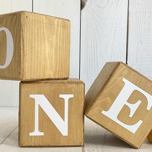 Puede incluir: Tres bloques de madera que deletrean la palabra "ONE" en letras blancas. Los bloques est&aacute;n apilados uno encima del otro, con el bloque "O" en la parte superior, el bloque "N" en el medio y el bloque "E" en la parte inferior.