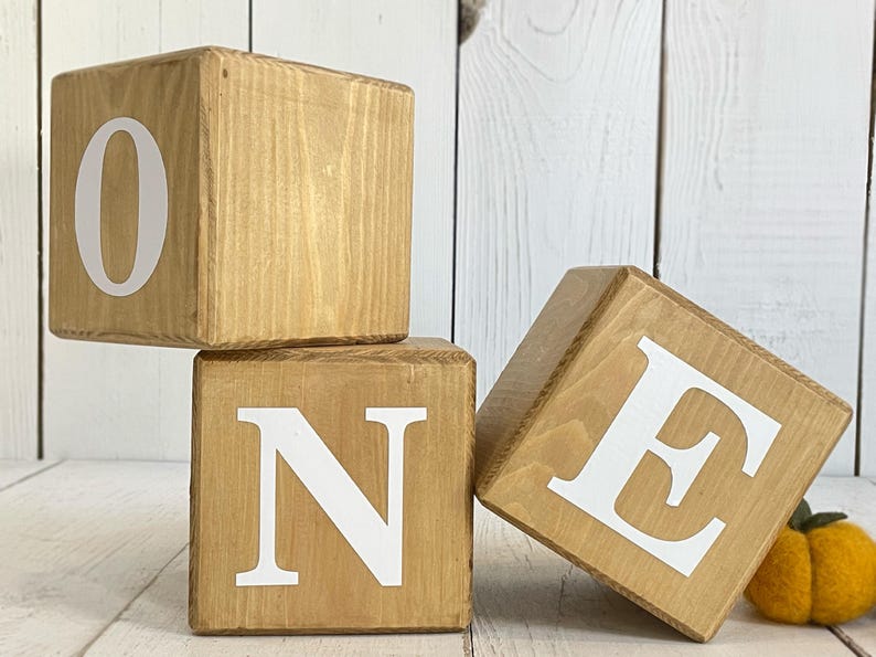 Puede incluir: Tres bloques de madera que deletrean la palabra "ONE" en letras blancas. Los bloques est&aacute;n apilados uno encima del otro, con el bloque "O" en la parte superior, el bloque "N" en el medio y el bloque "E" en la parte inferior.
