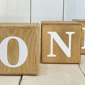 Puede incluir: Tres bloques de madera que deletrean la palabra "ONE" en letras blancas. Los bloques son de color marr&oacute;n claro y tienen un acabado r&uacute;stico.