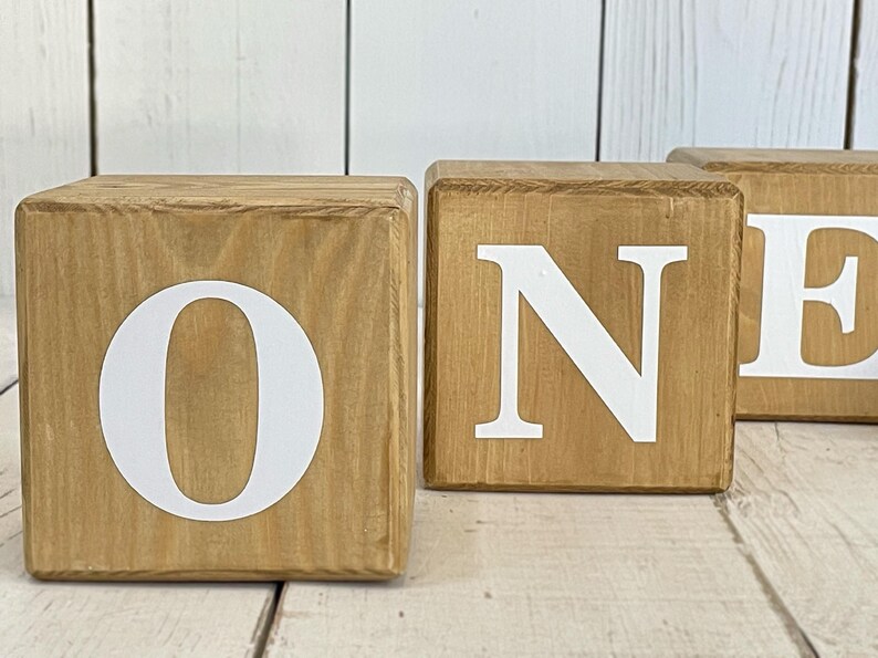 Puede incluir: Tres bloques de madera que deletrean la palabra "ONE" en letras blancas. Los bloques son de color marr&oacute;n claro y tienen un acabado r&uacute;stico.