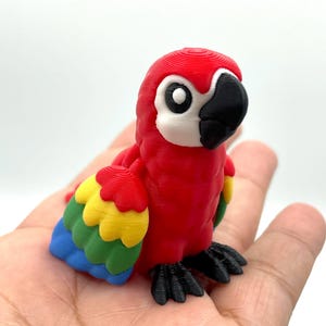 May include: A vibrant, 3D-printed parrot figurine. The bird features a red body, a black beak, and multi-colored wings with yellow, green, and blue feathers. The parrot is perched on a surface, showcasing its detailed design.