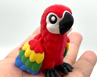 May include: A vibrant, 3D-printed parrot figurine. The bird features a red body, a black beak, and multi-colored wings with yellow, green, and blue feathers. The parrot is perched on a surface, showcasing its detailed design.