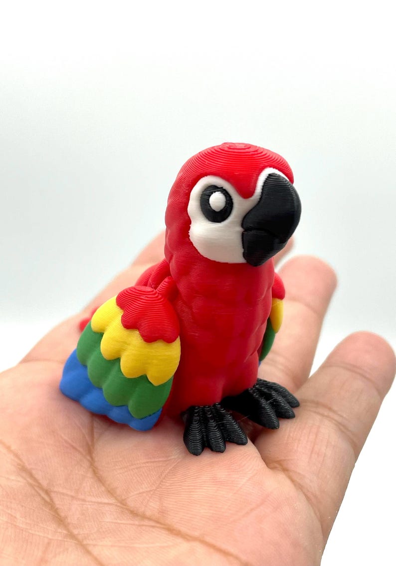 May include: A vibrant, 3D-printed parrot figurine. The bird features a red body, a black beak, and multi-colored wings with yellow, green, and blue feathers. The parrot is perched on a surface, showcasing its detailed design.
