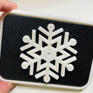 May include: A white snowflake-shaped button with a black background. The button is made of plastic and has a raised design.