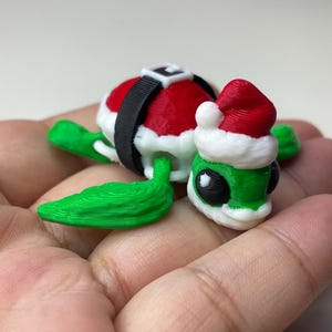 May include: A small, green and red 3D printed turtle wearing a Santa hat and a black belt. The turtle is sitting on a hand.