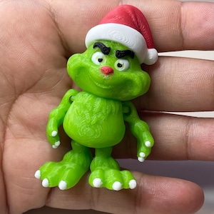 May include: A green figurine of a character wearing a red and white Santa hat. The figurine has a grumpy expression with black eyebrows and a small smile. The feet and hands have white tips.