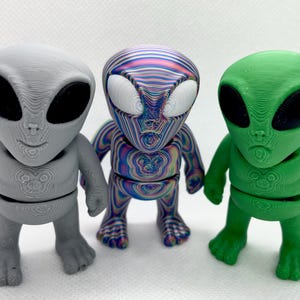 May include: Three alien figurines in gray, multicolored, and green. Each has large black eyes and a small mouth. The gray alien is solid, the multicolored one has a swirling pattern, and the green one is a solid color. The figurines are standing.