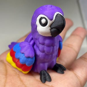 May include: A 3D-printed purple parrot figurine with a white face, black beak, and colorful wings. The wings feature red, yellow, and blue accents. The parrot's feet are black.