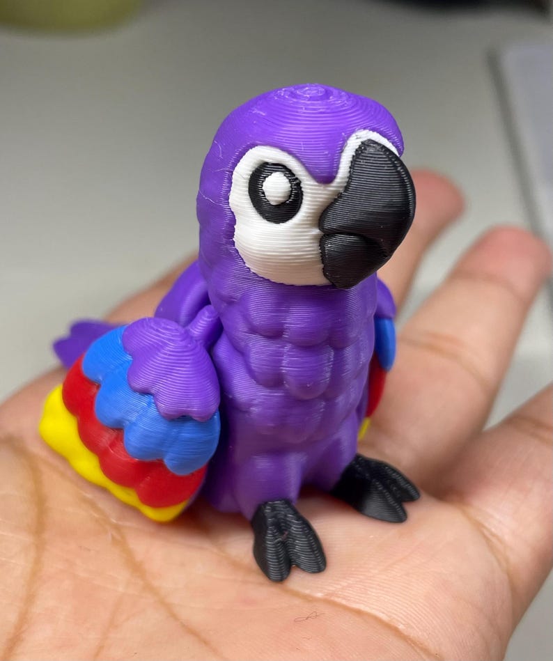 May include: A 3D-printed purple parrot figurine with a white face, black beak, and colorful wings. The wings feature red, yellow, and blue accents. The parrot's feet are black.