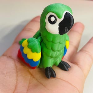 May include: A 3D-printed green parrot figurine with a white face, black beak, and large black eyes. The parrot's wings are detailed with red, yellow, and blue accents. The figurine is held in a hand, showcasing its size and detail.