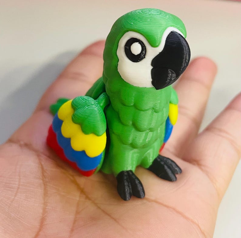 May include: A 3D-printed green parrot figurine with a white face, black beak, and large black eyes. The parrot's wings are detailed with red, yellow, and blue accents. The figurine is held in a hand, showcasing its size and detail.