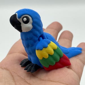 May include: A blue parrot figurine with a black beak and feet, and a white eye. The parrot's wing is a mix of red, yellow, and green. The figurine is resting on a hand.