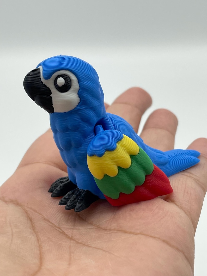 May include: A blue parrot figurine with a black beak and feet, and a white eye. The parrot's wing is a mix of red, yellow, and green. The figurine is resting on a hand.
