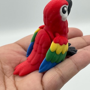 May include: A vibrant, 3D-printed parrot figurine. The bird features a bright red body, with wings in shades of yellow, green, and blue. The parrot's head has a white face, black beak, and black feet. The figurine is a decorative item.