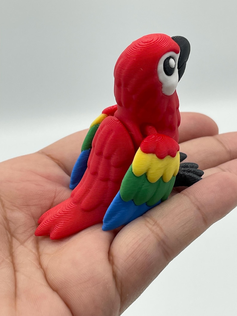 May include: A vibrant, 3D-printed parrot figurine. The bird features a bright red body, with wings in shades of yellow, green, and blue. The parrot's head has a white face, black beak, and black feet. The figurine is a decorative item.