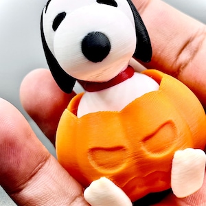May include: A 3D-printed Snoopy figurine emerging from an orange pumpkin. The Snoopy figure has a white head with black ears and a black nose, wearing a red collar. The pumpkin has carved-out eyes and a smiling mouth.