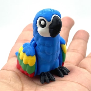 May include: A blue parrot figurine with a white face, black beak, and black feet. The parrot has yellow, red, and green accents on its wings. The figurine is held in a person's hand.