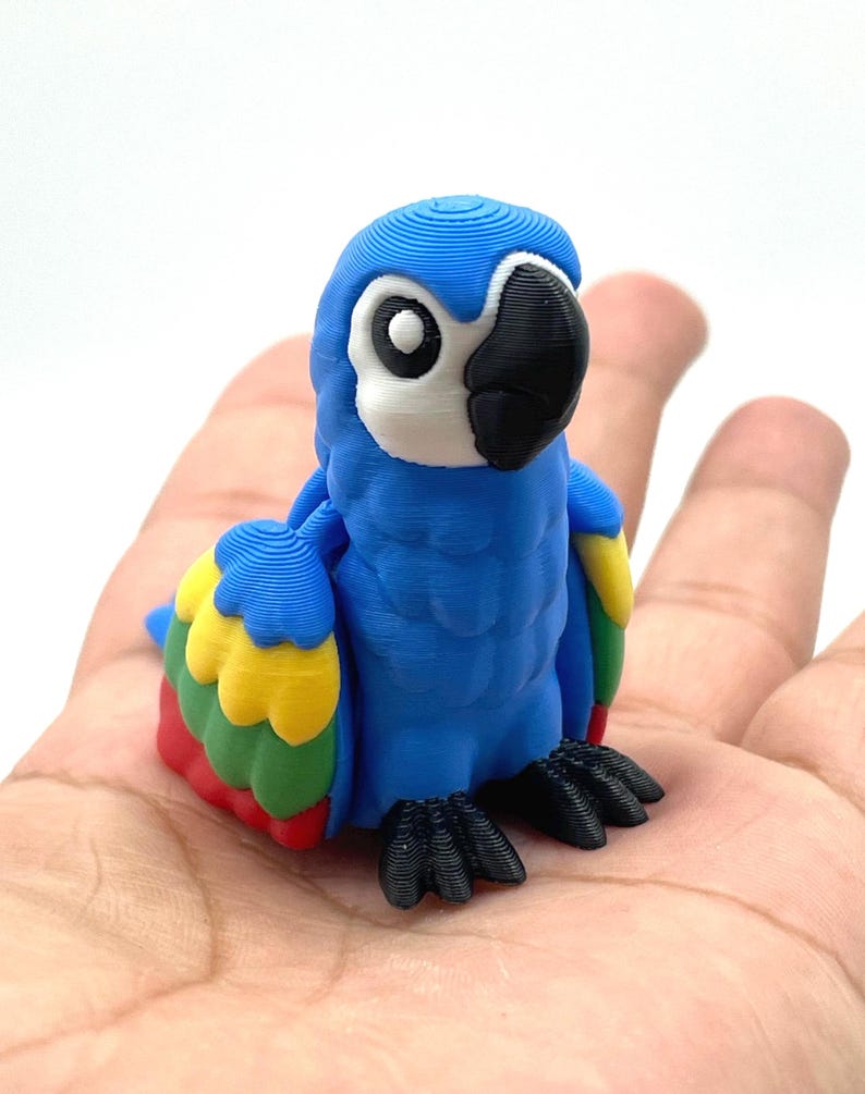 May include: A blue parrot figurine with a white face, black beak, and black feet. The parrot has yellow, red, and green accents on its wings. The figurine is held in a person's hand.