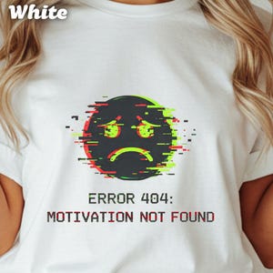 May include: White t-shirt featuring a distressed graphic of a sad emoji with red and green glitch effects. Below the emoji, the text reads "ERROR 404: MOTIVATION NOT FOUND" in a pixelated font.
