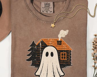 May include: Espresso-colored t-shirt with a graphic of a ghost in front of a cabin with a smoking chimney. The shirt has a Comfort Colors tag and a gold star necklace. The shirt is a size XL.