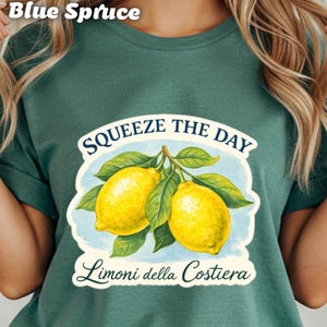 May include: A teal t-shirt featuring a graphic of two yellow lemons with green leaves. The text "Squeeze the Day" arches above the lemons, with "Limoni della Costiera" below. The words "Blue Spruce" are at the top.