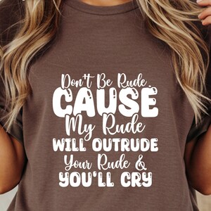 May include: A brown t-shirt with the white text "Don't Be Rude... CAUSE My Rude Will Outrude Your Rude & YOU'LL CRY". The shirt has short sleeves and a crew neck.