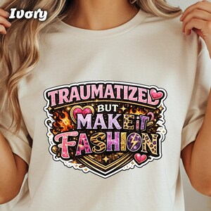 May include: Ivory-colored t-shirt featuring a colorful graphic that reads "TRAUMATIZE BUT MAKE IT FASHION." The design includes hearts, flames, and a lightning bolt. The text is in a mix of pink, purple, and gold colors.