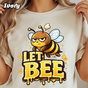 May include: Ivory-colored t-shirt featuring a cartoon bee with an angry expression. The bee is brown and yellow with blue wings. The text "LET BEE" is in yellow with a dripping effect.