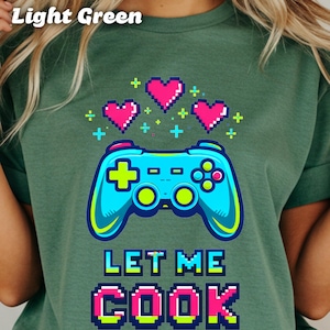 May include: Light green t-shirt featuring a pixelated design. The design includes a blue video game controller, pink pixel hearts, and the text "LET ME COOK" in a pixelated font. The shirt is a solid light green color.