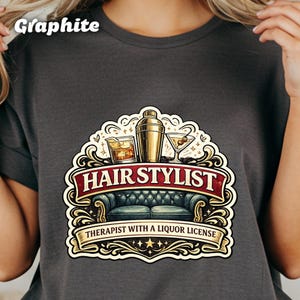 May include: Graphite gray t-shirt featuring a decorative design with a cocktail shaker, two cocktail glasses, and a whiskey glass. The design includes the text "HAIR STYLIST" and "THERAPIST WITH A LIQUOR LICENSE" in a banner.