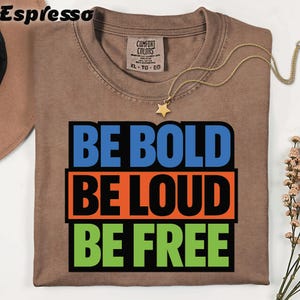 May include: A brown t-shirt with the words "BE BOLD BE LOUD BE FREE" in blue, orange, and green block letters. A gold star pendant necklace and a Comfort Colors tag are visible. The shirt is folded.