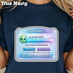 May include: Navy blue t-shirt featuring a graphic of a loading screen with the text "LOADING..." and "ADULTING IN PROGRESS." The graphic includes a sad alien face and the options "Refresh" and "Go Outside."