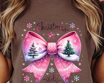 May include: Brown t-shirt featuring a large pink Christmas bow with a winter scene and two Christmas trees. The words "Christmas" and "girly" are written in cursive script, surrounded by snowflakes.